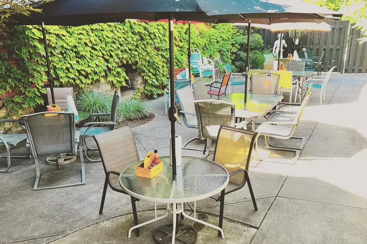 Rizz's patio on a bright summer afternoon, the existing dark patio umbrellas open, a small round table in the foreground, the ivy wall in deep green behind the seating area. Ready to host a casual team dinner with no extra dressing required.